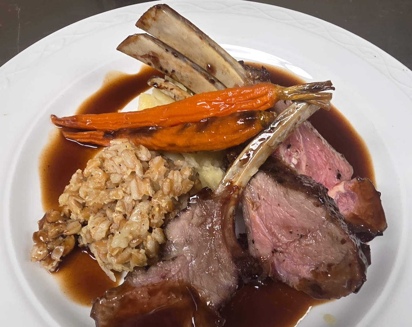 A rack of lamb with roasted carrots, mashed potatoes, a grain side, and a rich brown jus.